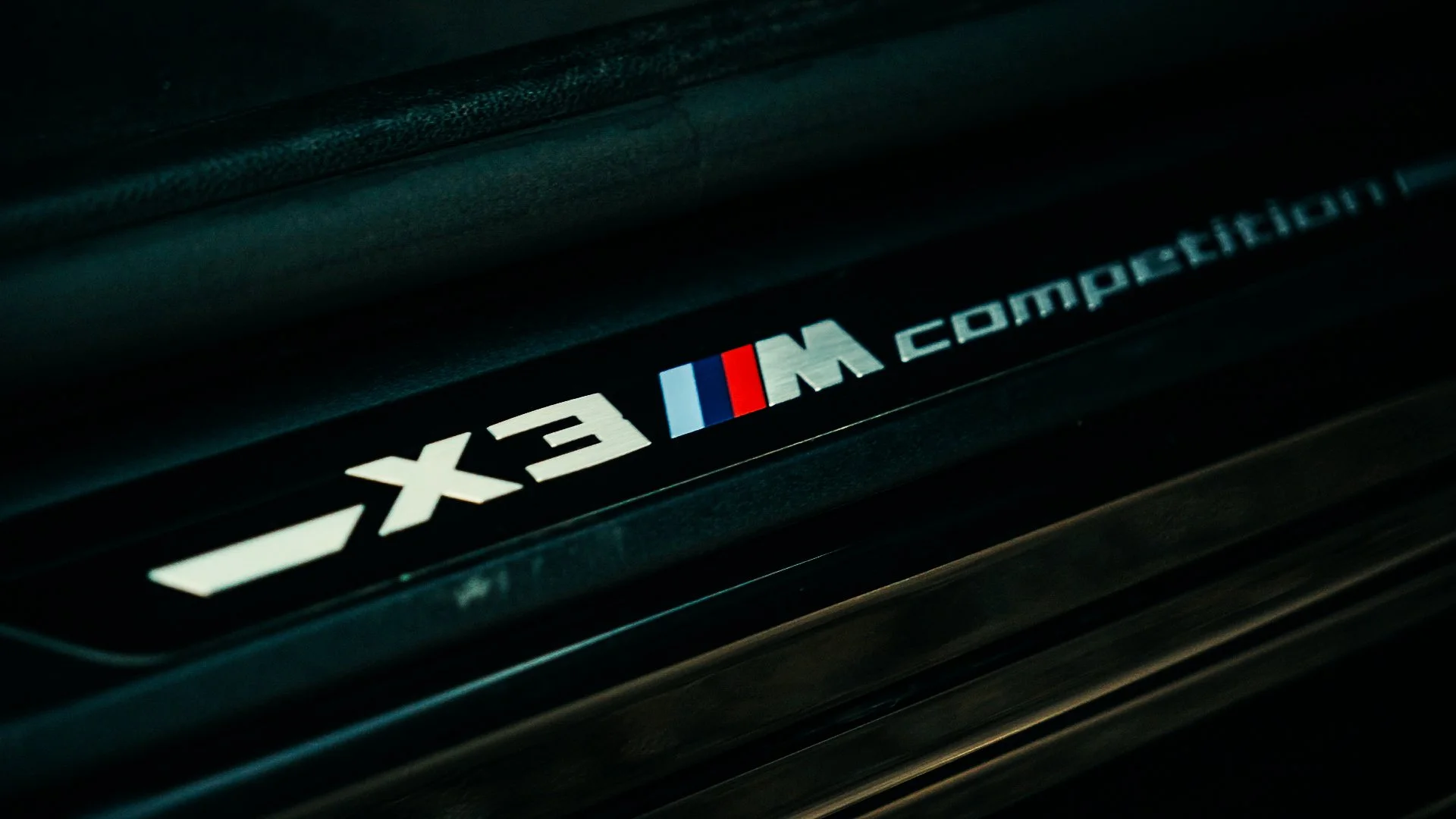 Close-up of a BMW M Series emblem on a car, showing the letters 'X3 M' with the BMW M logo in blue, purple, and red stripes.
