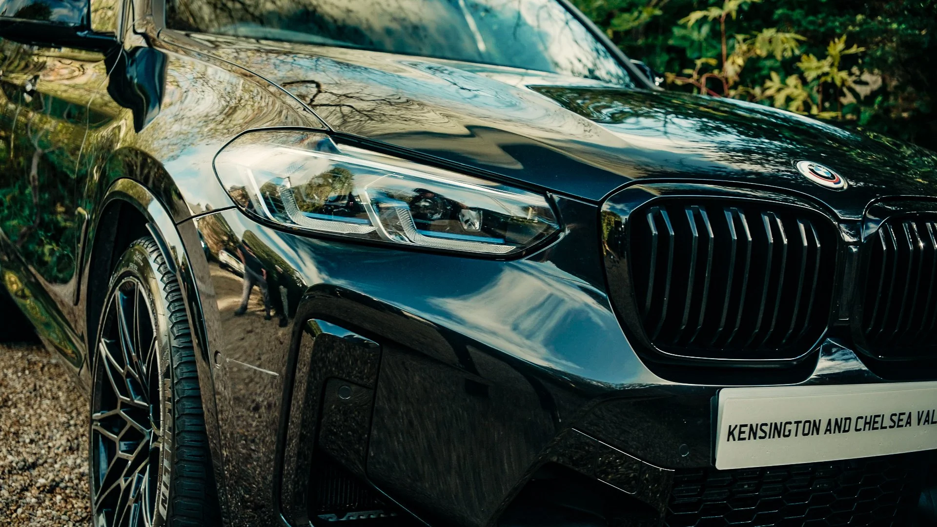 Close-up of a black luxury BMW car with reflections on its shiny surface, parked outdoors on gravel with green foliage in the background.
