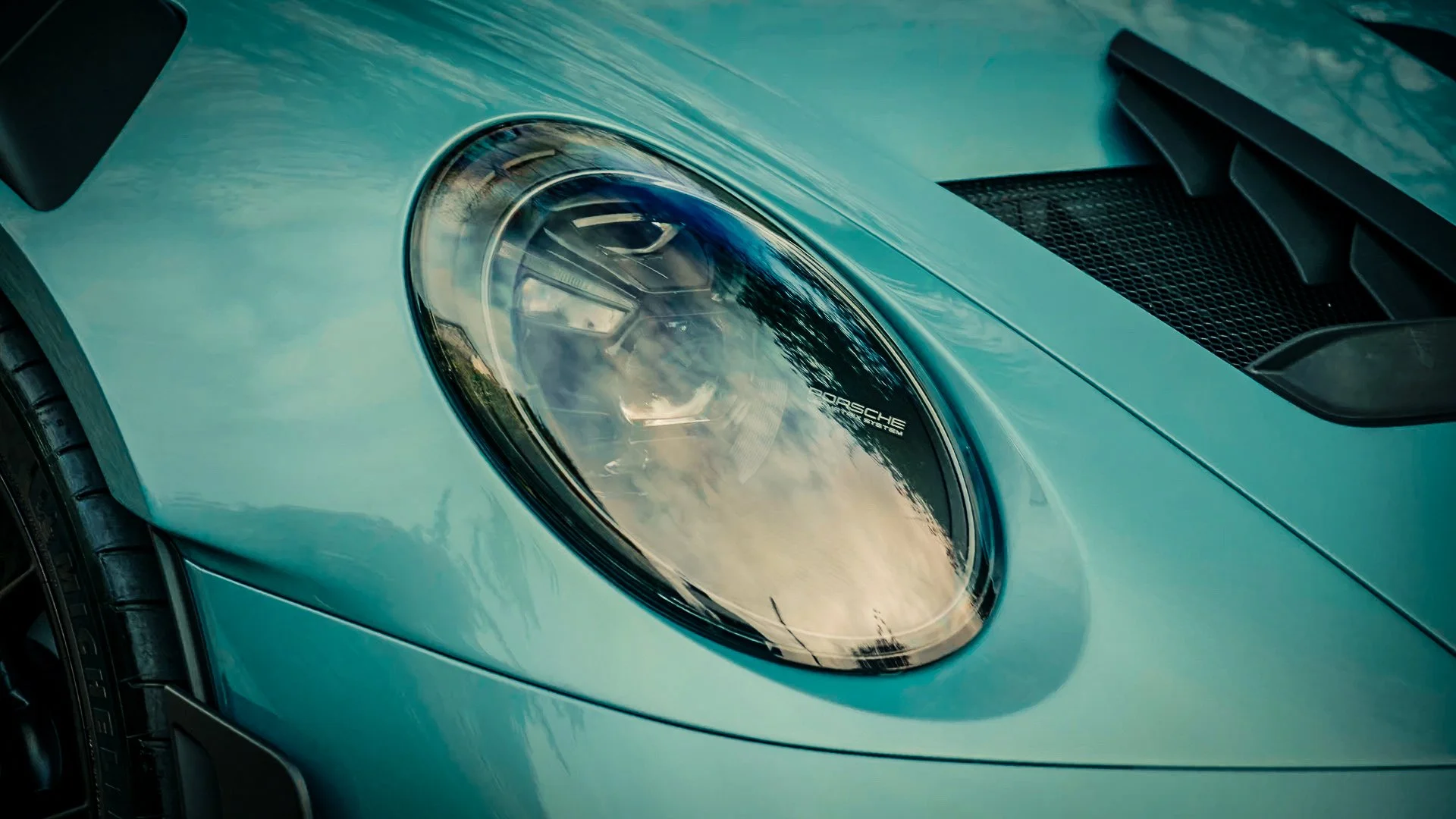 Close-up of a light blue Porsche car's front headlight and hood, with a ventilation grille nearby.