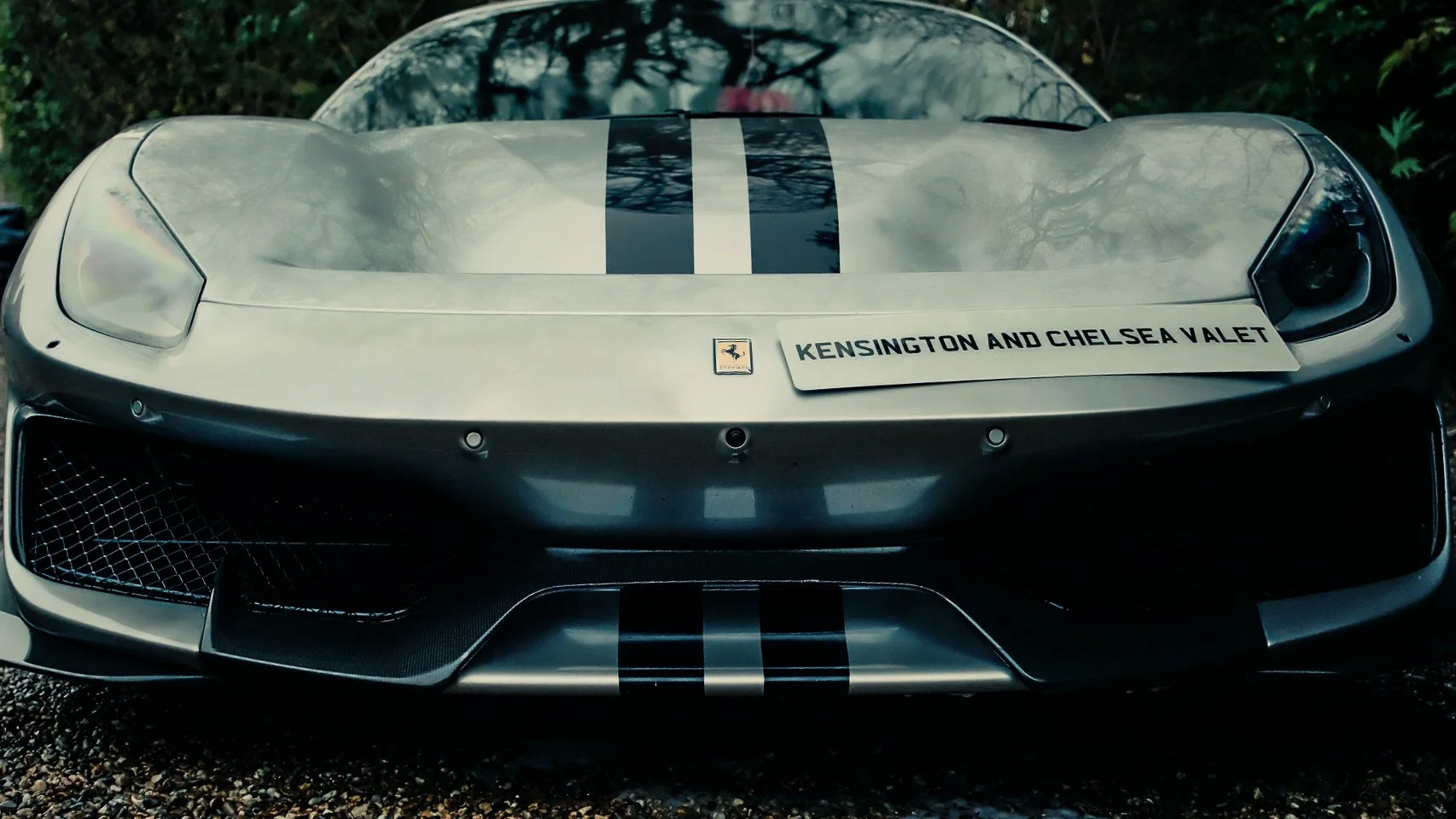 Front view of a silver Ferrari sports car with black racing stripes, parked outdoors with trees reflected on the windshield, and a sign on the hood that says 'Kensington and Chelsea Valet'.