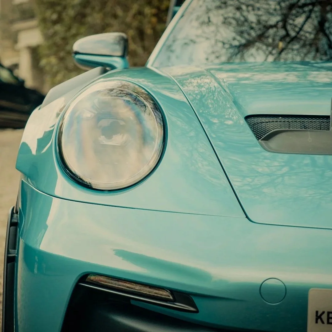 Close-up of the front left side of a light blue sports car, showing the round headlight, a side mirror, and part of the hood with a vent.
