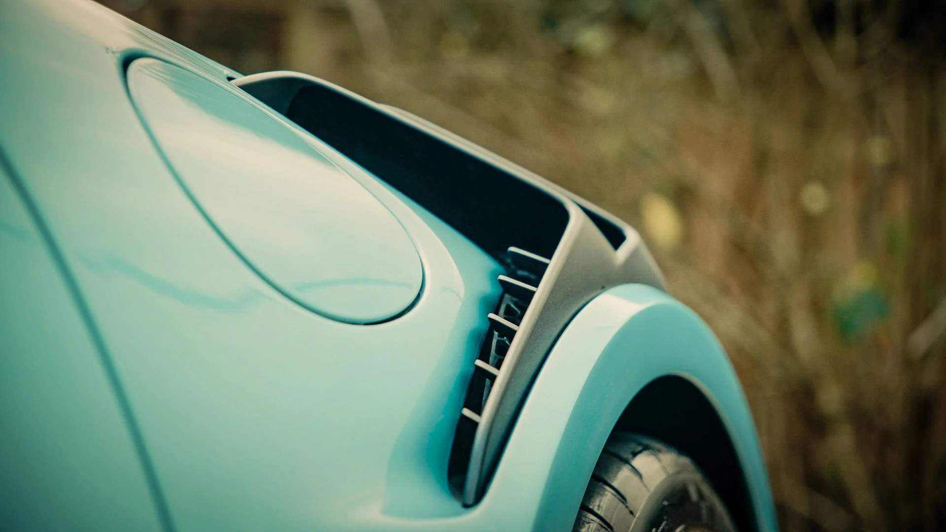 Close-up of a vintage teal-colored car's front section with a focus on the round hood vent and the wheel, set against a blurred natural background.