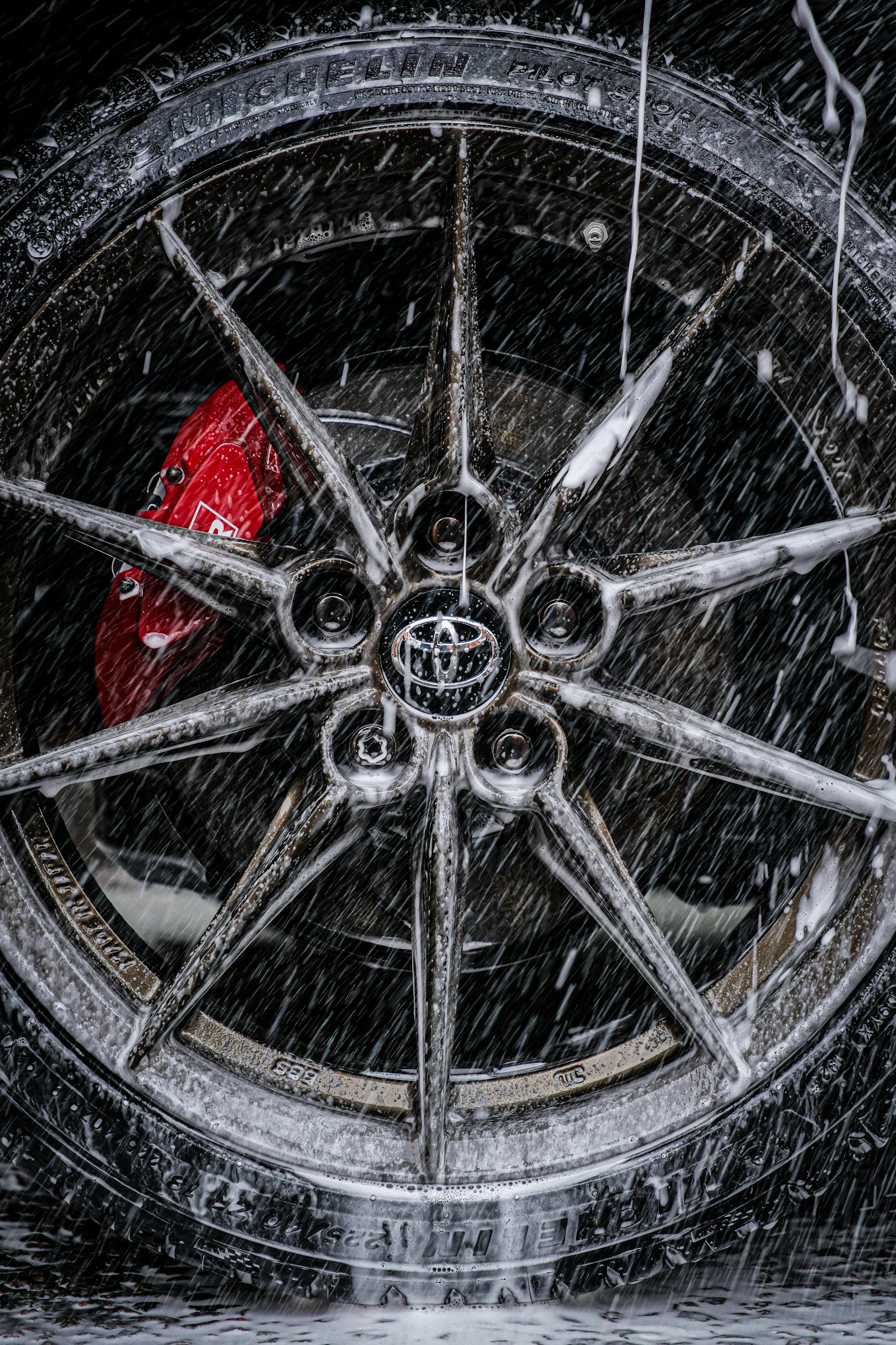 A close-up of a car wheel with soap suds and water, showing the Toyota logo at the center, with red brake calipers visible through the spokes.