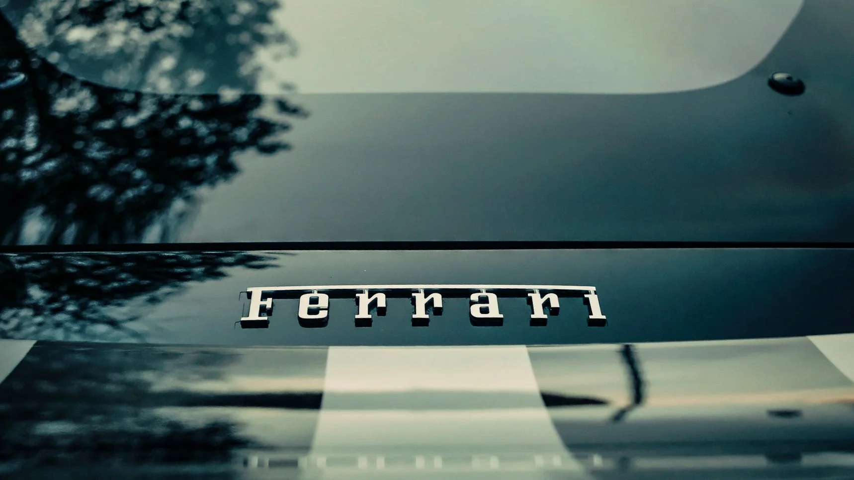 Close-up of a black car's hood with a chrome Ferrari emblem and reflections of trees and the sky.