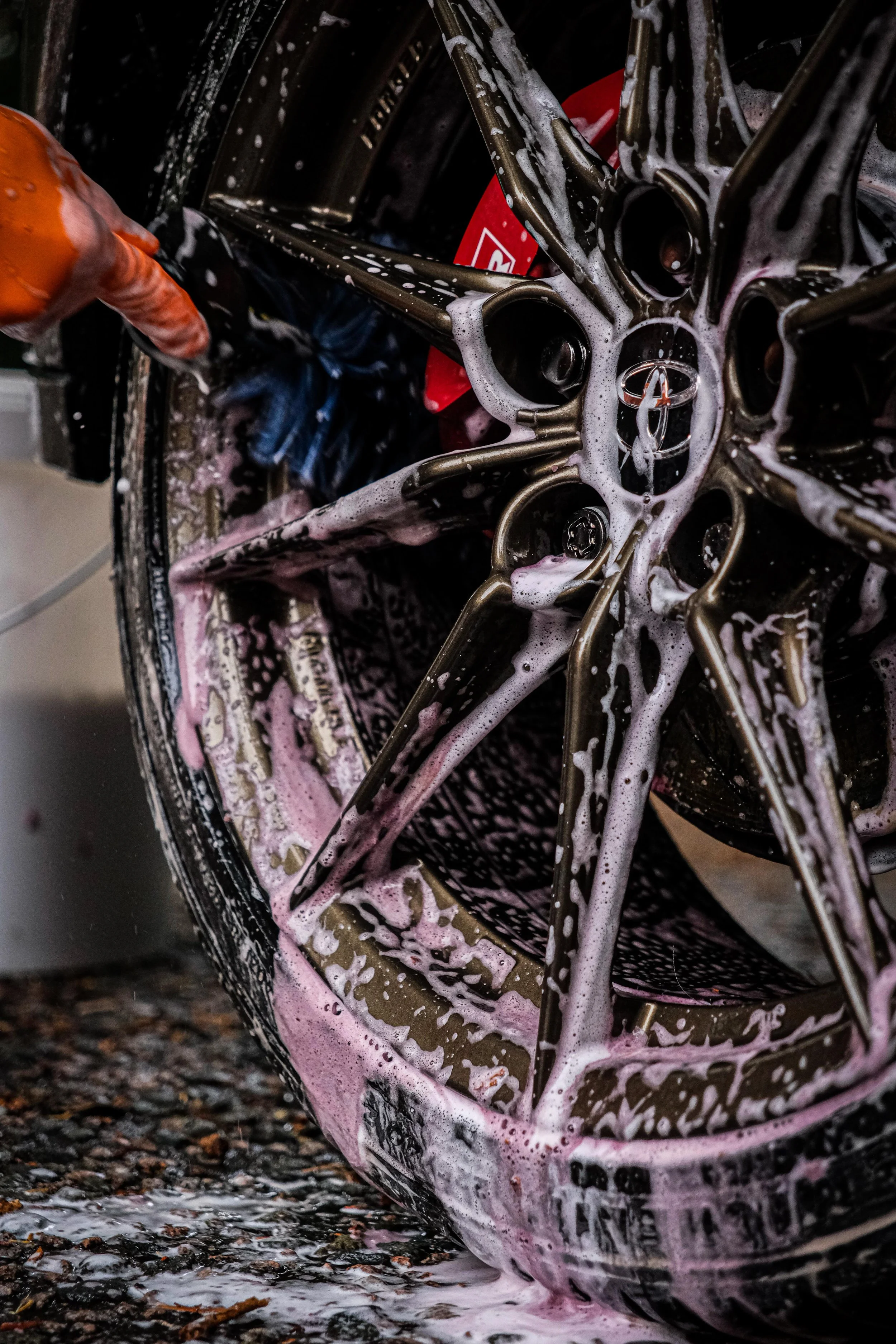 A person is washing a car wheel with soap suds, using a brush or sponge, on a gravel surface.