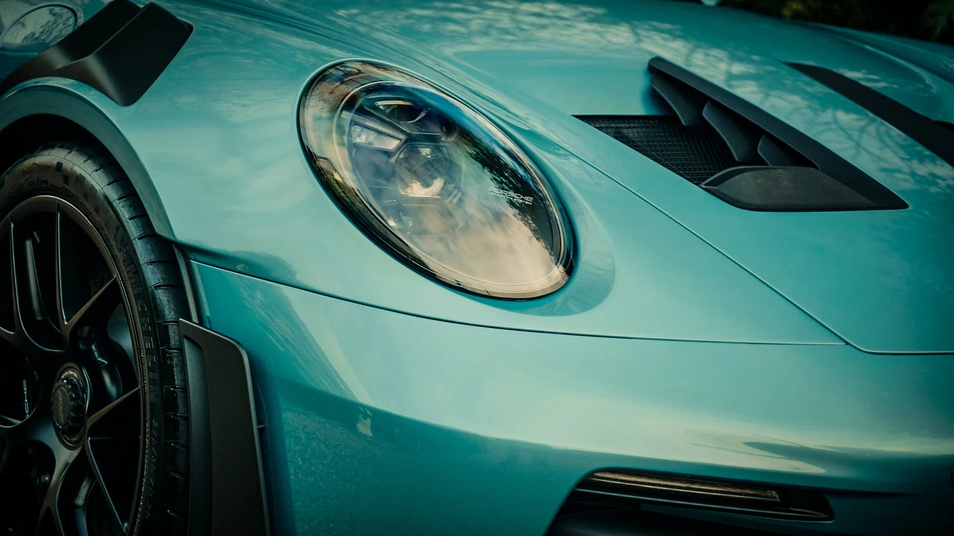 Close-up of the front part of a light blue Porsche sports car, showing the headlight, part of the hood, and a black vent.