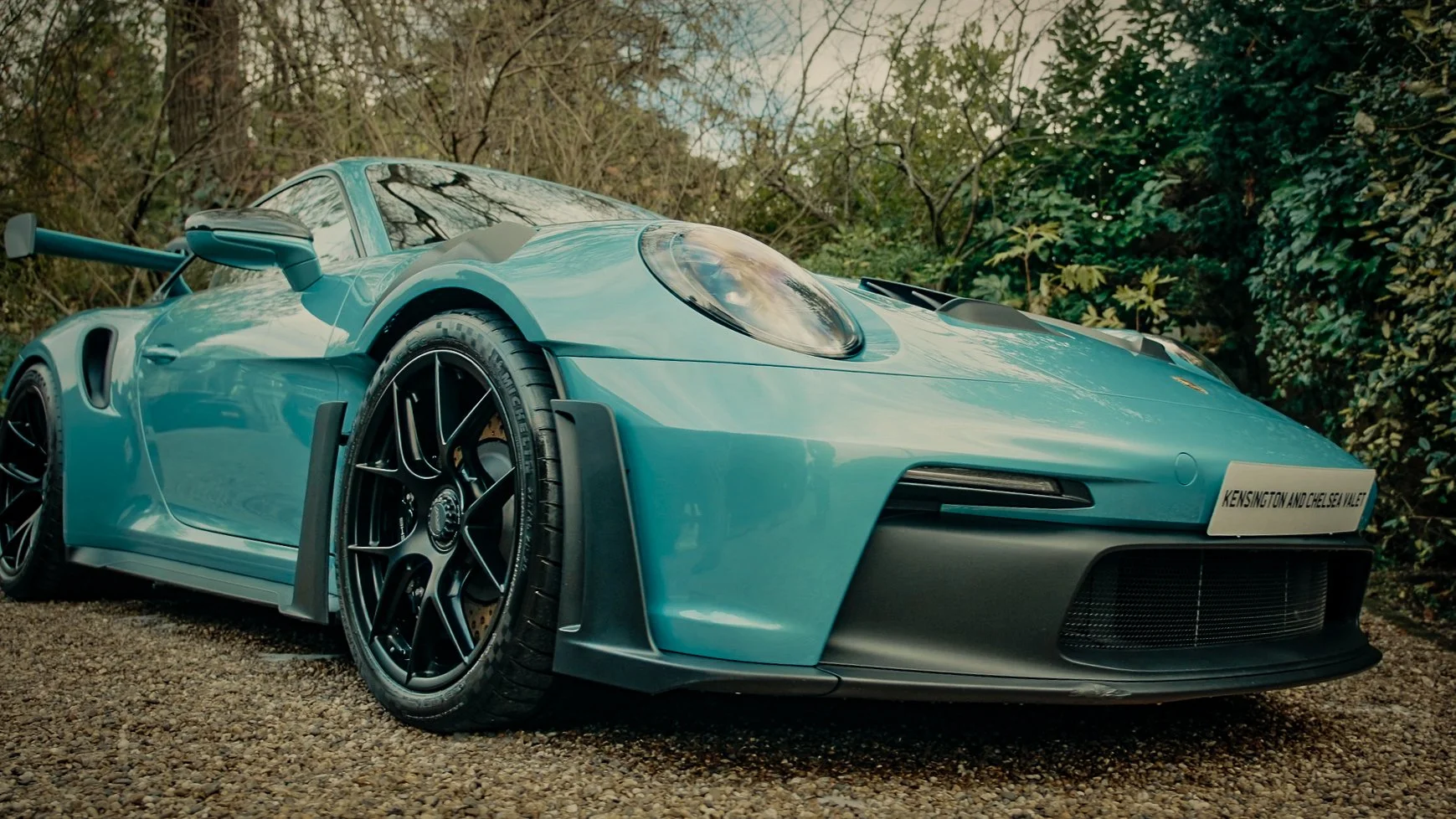 A light blue Porsche 911 GT3 racing car parked on a gravel surface, with a black front grille, black wheels, and a rear wing, surrounded by trees