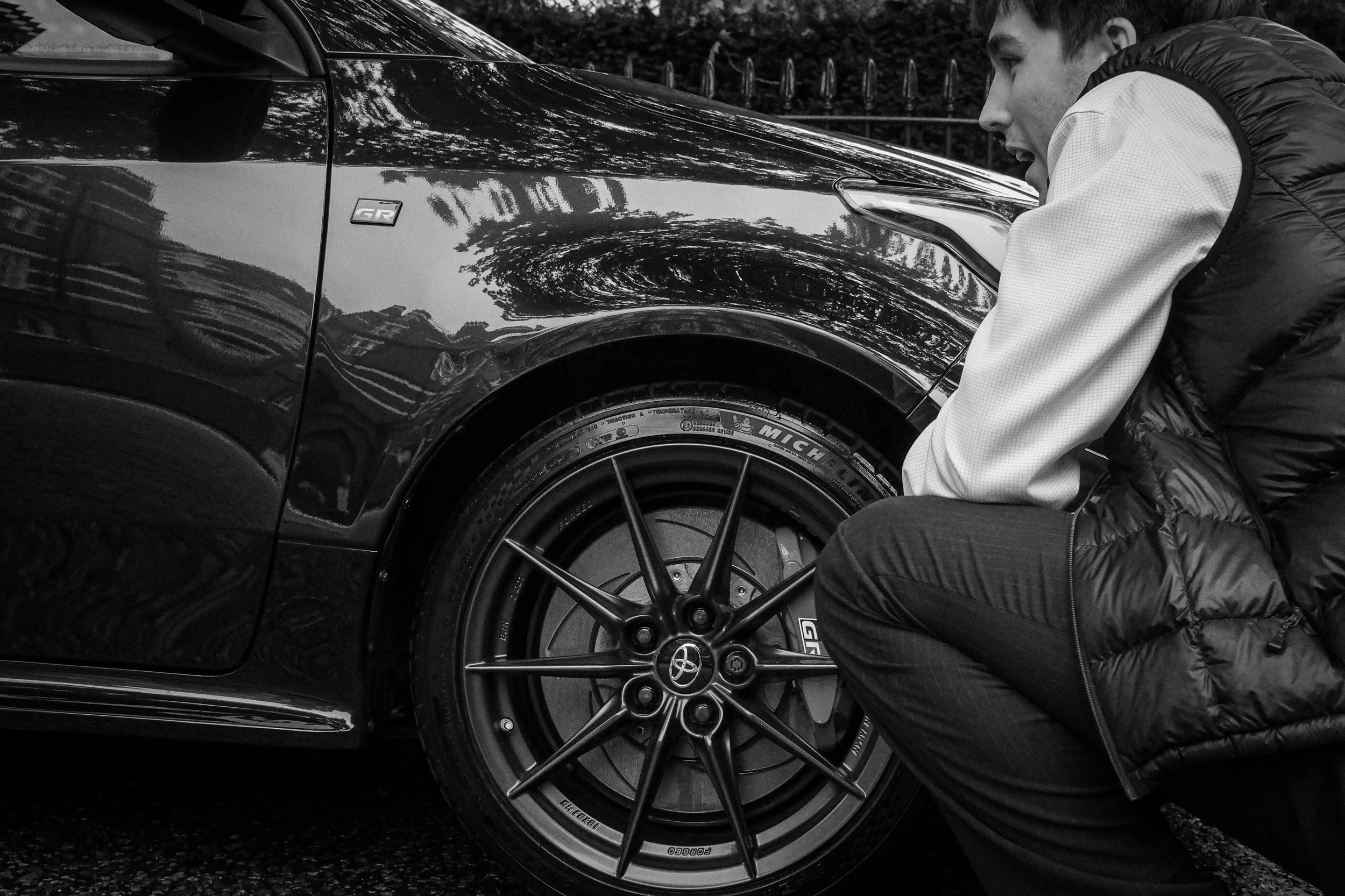 A man crouching beside a black sports car, inspecting the front wheel."