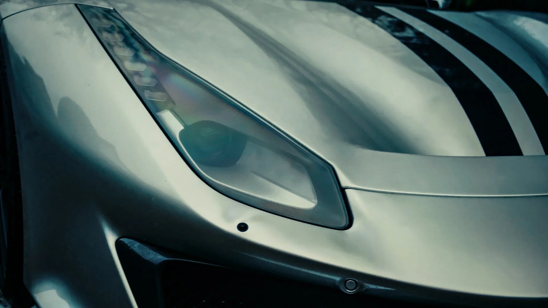Close-up of front part of a silver sports car, showing the sleek headlight and part of the hood with black racing stripes.