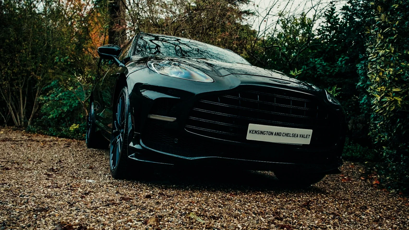 A black luxury sports car parked on a gravel path surrounded by trees and bushes, with a sign on the front plate that reads "Kensington and Chelsea Valet."