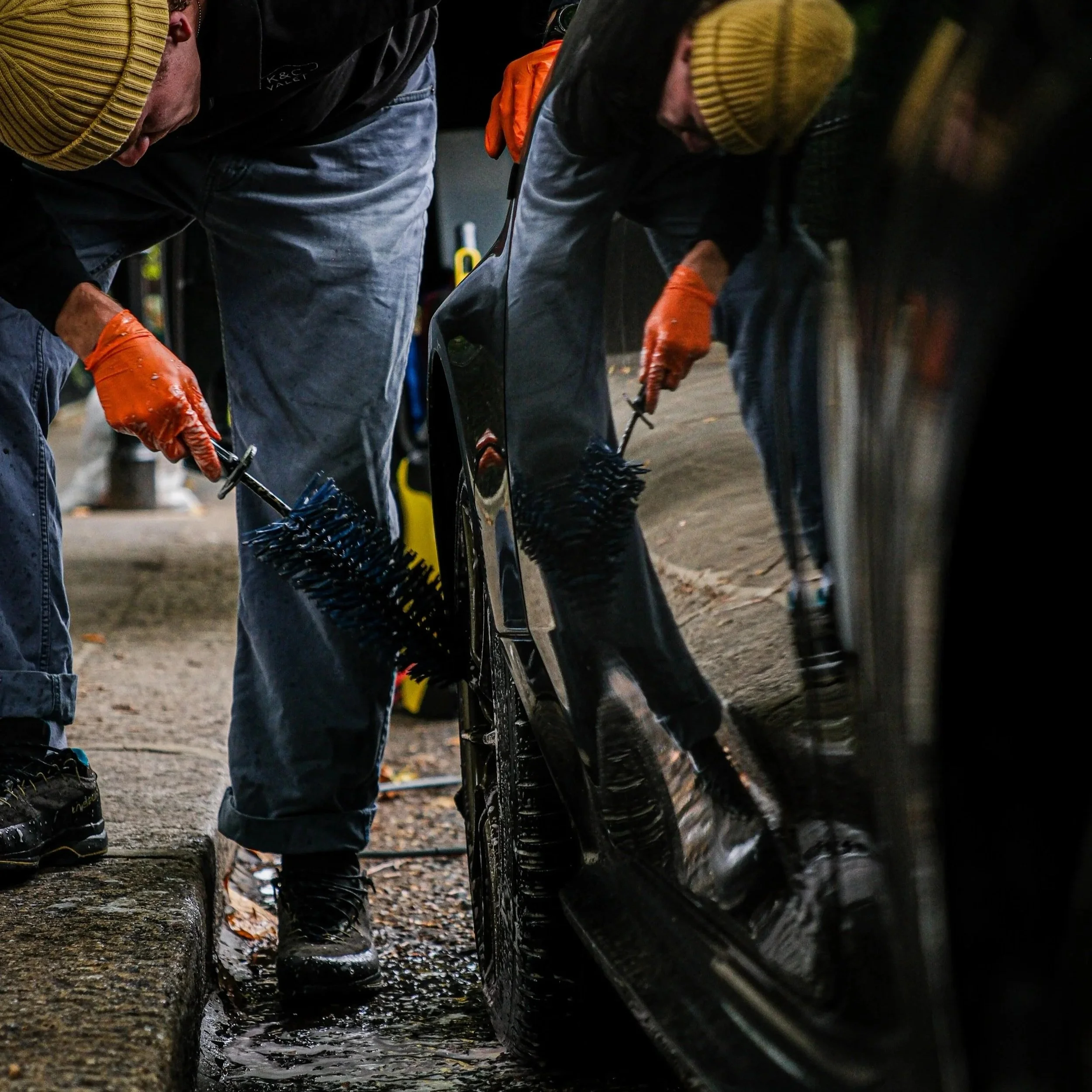 Two people washing a black car with brushes while standing on the street, wearing gloves and warm clothing.