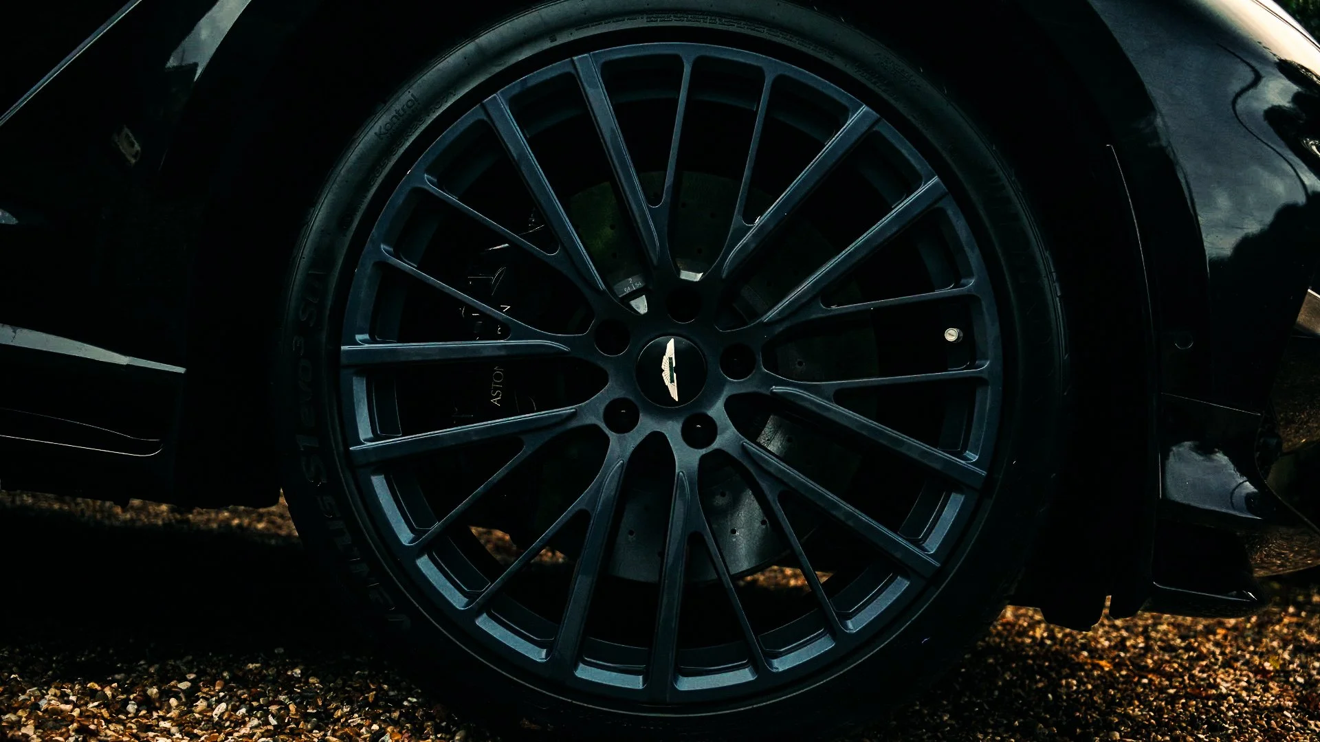 Close-up of a black car wheel with a multi-spoke alloy rim and a visible Aston Martin logo in the center, parked on a gravel surface.