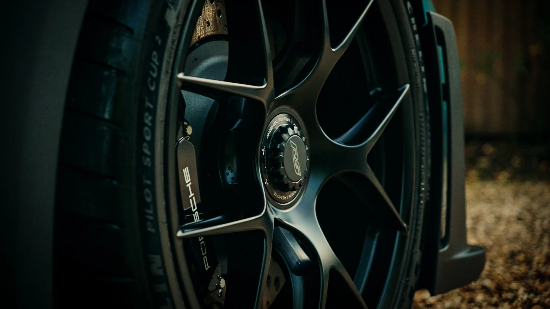 Close-up of a black alloy wheel with the Porsche and RS logos, mounted on a tire, outdoors with a blurred background.