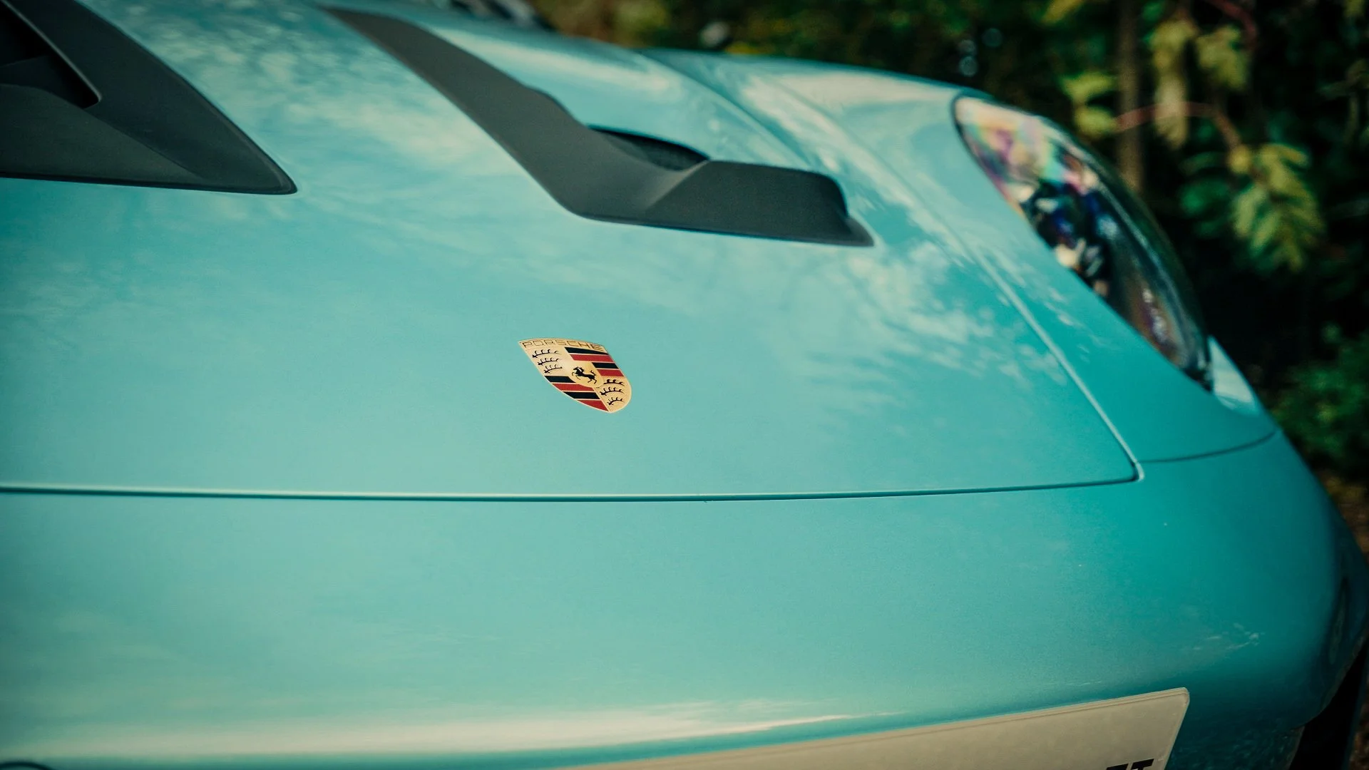 Close-up of the front hood of a teal Porsche sports car, showing the Porsche emblem and headlight, with a blurred green foliage background.