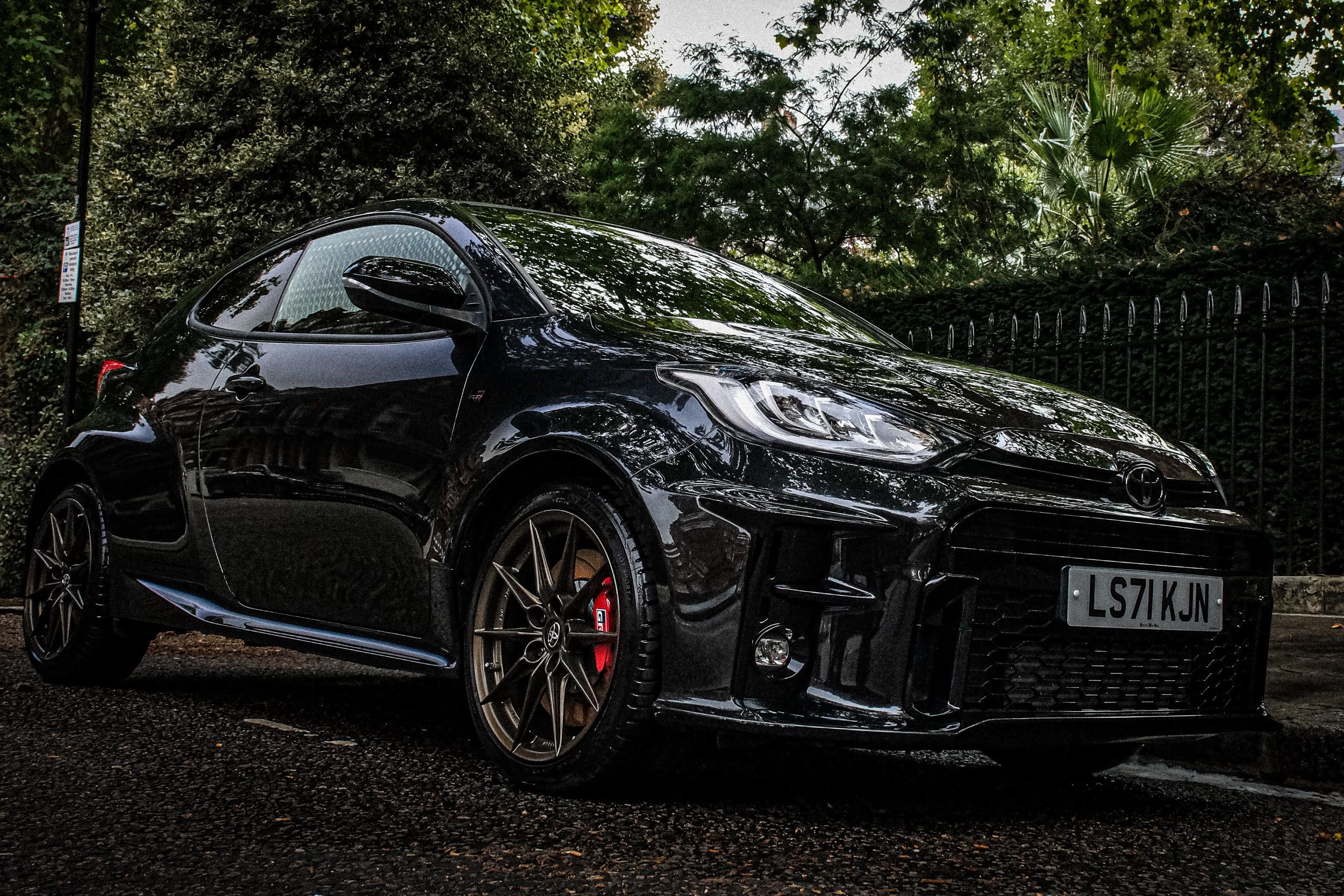 Black sports car parked on a dimly lit street with trees and a black fence in the background.