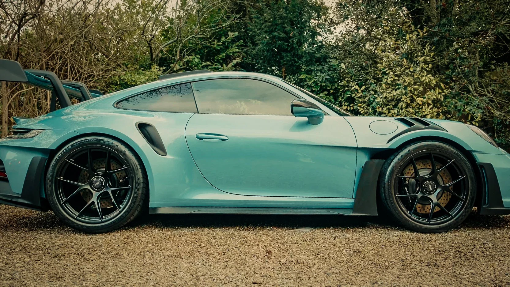 A sleek, light blue sports car with black wheels parked on gravel in front of green foliage.