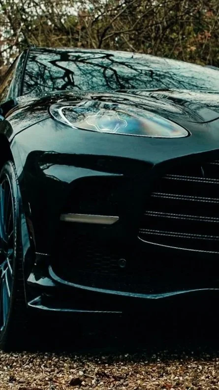 Close-up of the front end of a black luxury car, showing the headlight, front grille, and part of the hood, with leafless trees in the background.