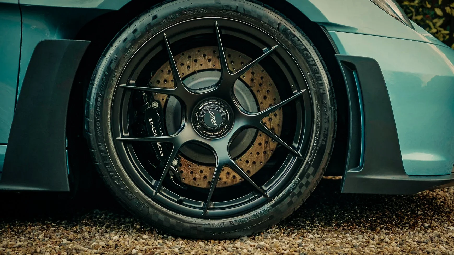 Close-up of a sports car wheel featuring a brake caliper labeled 'Porsche' and a drilled brake disc, with part of the car's body and ground visible.