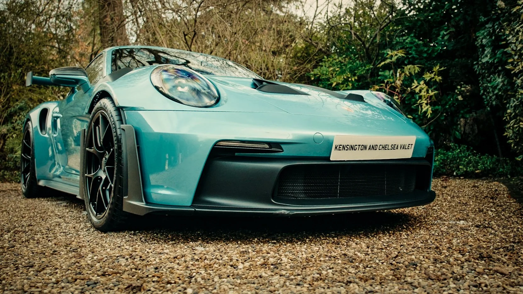 A blue sports car parked on gravel with trees and bushes in the background.