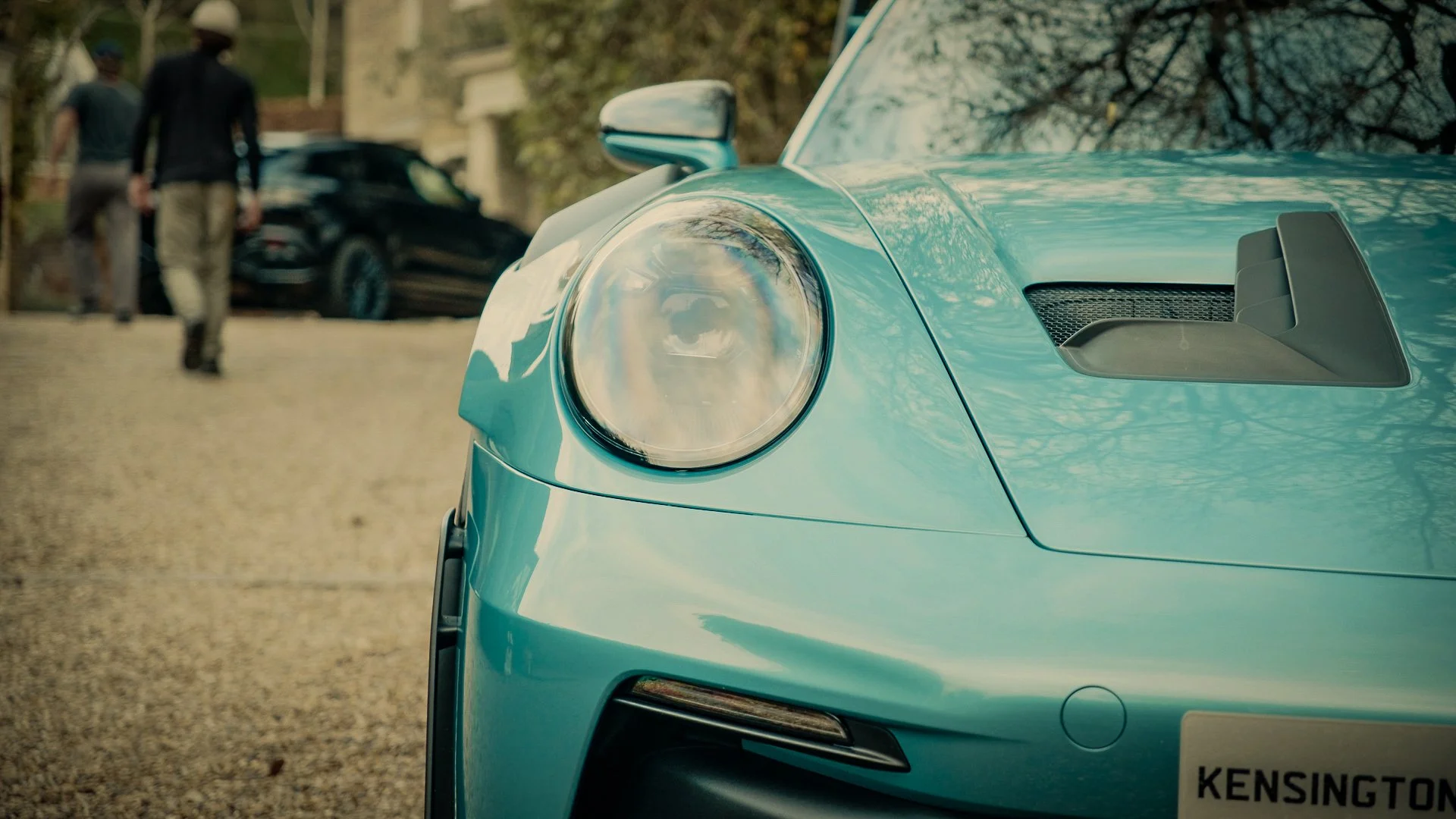 Close-up of the front of a teal sports car, showing the headlight, air intake, and part of the windshield with reflections of trees.