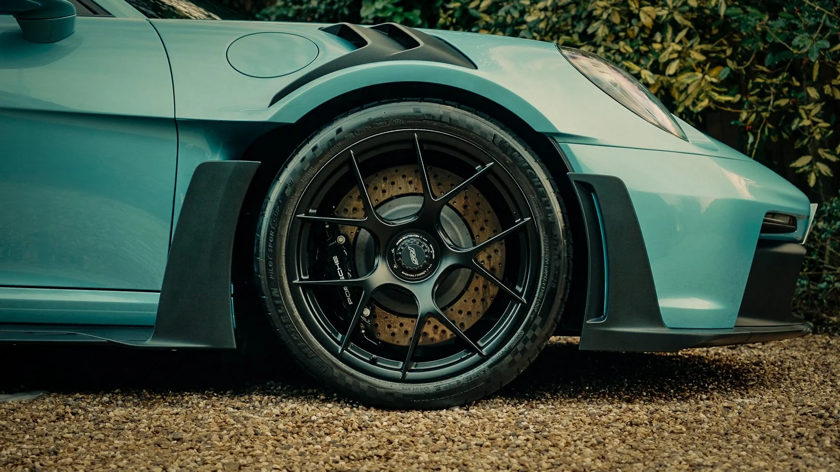 Close-up of a sports car's front right wheel, showcasing a black multi-spoke rim, a brake disc with drilled holes, and a tire on a gravel surface with greenery in the background.