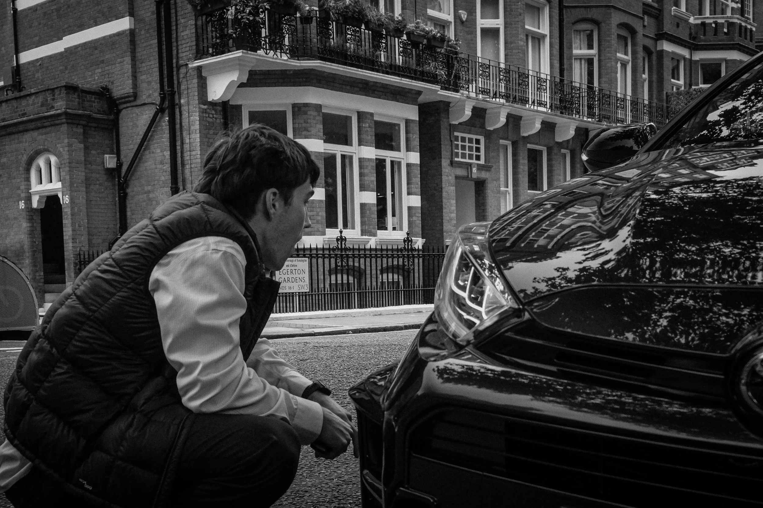 A man crouches next to a car parked on a city street, inspecting or working on the front of the vehicle, with residential buildings in the background.