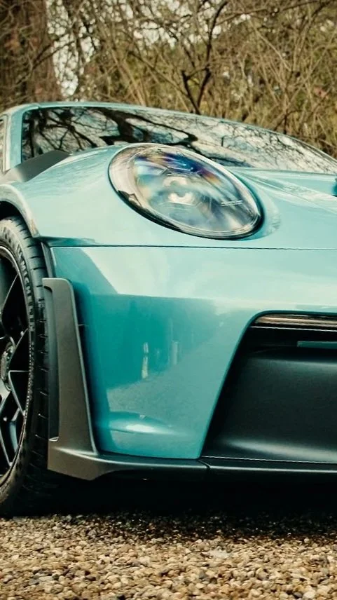 Close-up of the front left side of a turquoise sports car with black wheels and a foggy daytime background of trees.