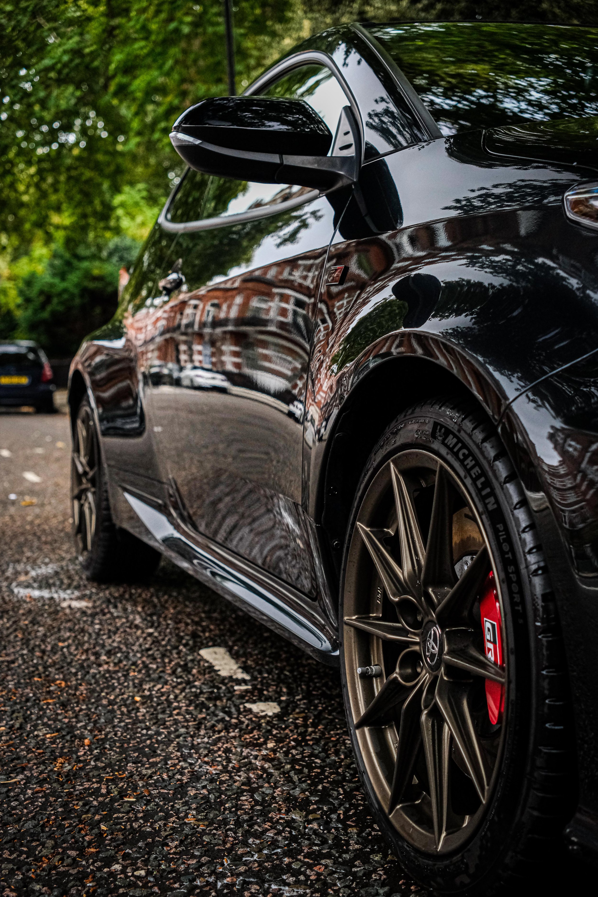 Close-up of a black sports car with gold rims parked on a street with trees and buildings reflected on its surface.