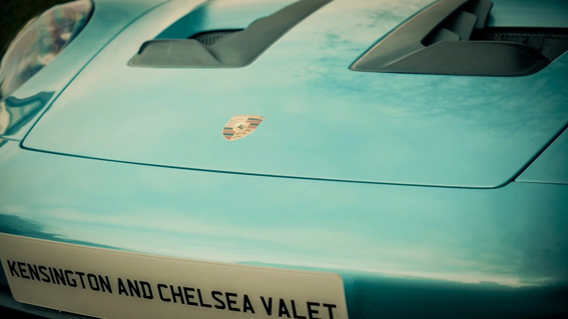 Close-up of a turquoise Porsche car with a Porsche emblem on the hood and a sign that reads "Kensington and Chelsea V Alet."