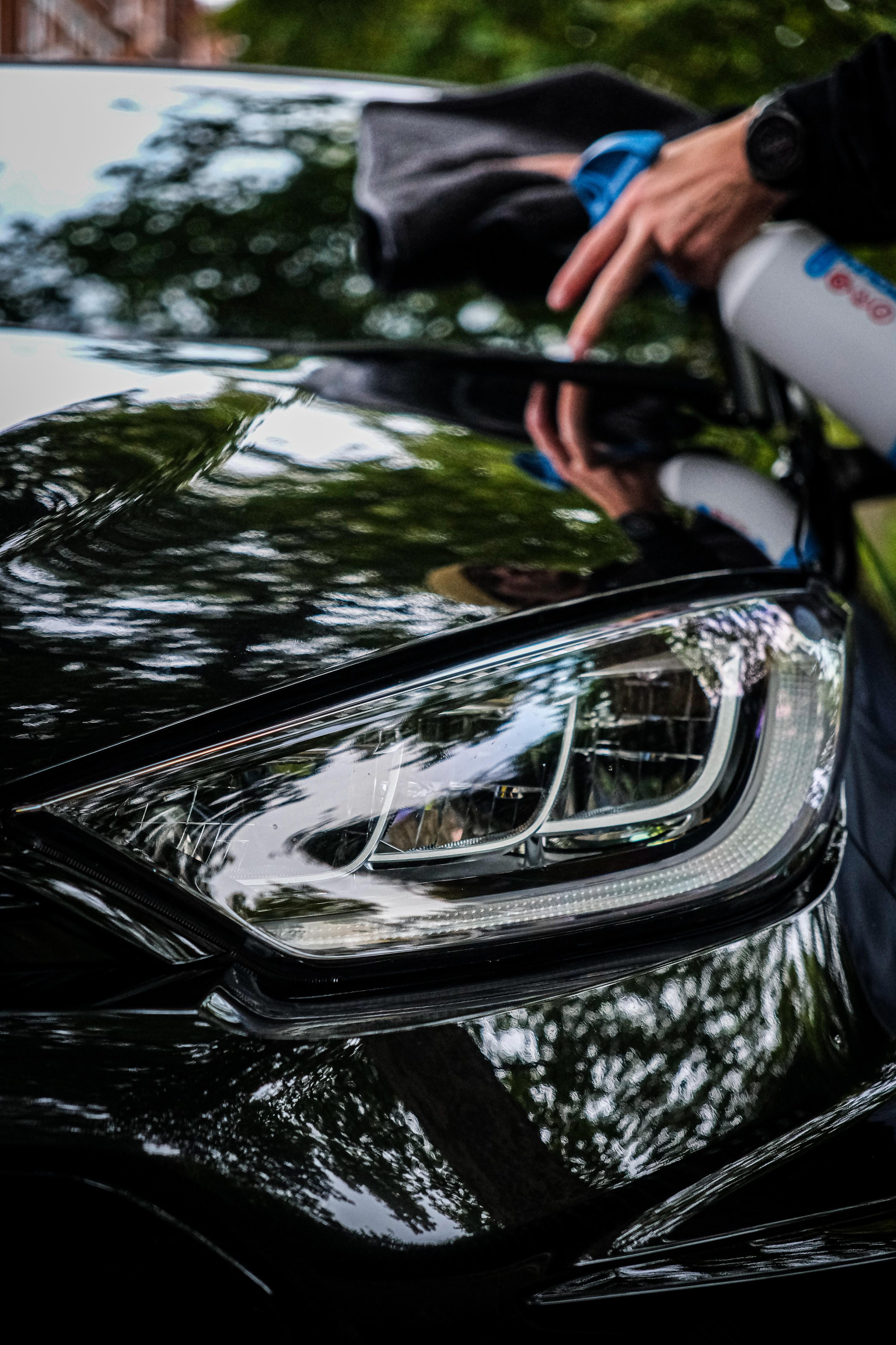 Close-up of a black car's headlight reflecting greenery and a person with a grooming tool.