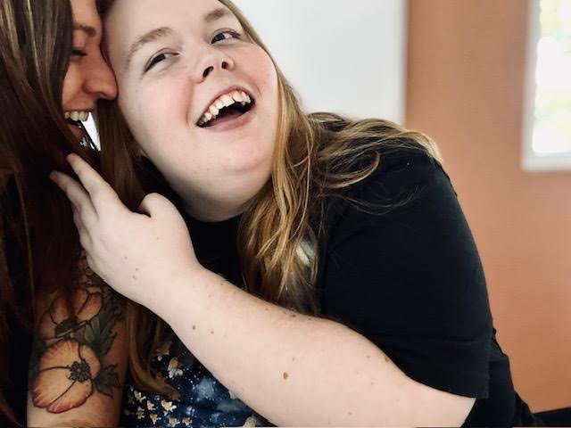Two young women smiling and close together, one with her hand on the other's neck, in an indoor setting with a window in the background.