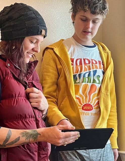 A young woman and a young man looking at a tablet together in a room with beige walls. The woman is wearing a black beanie and a red jacket, and the man is wearing a yellow hoodie and a white T-shirt with colorful text.
