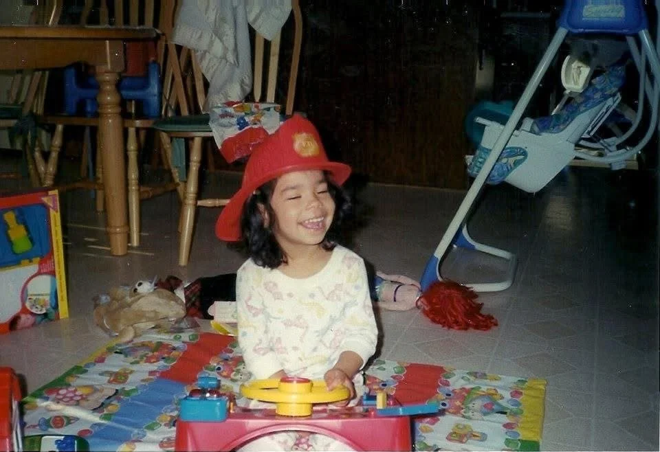 A young girl with shoulder-length dark hair smiling while playing with a toy keyboard, wearing a red firefighter hat. She is seated on a colorful mat surrounded by toys in a home setting with wooden chairs, a high chair, and scattered items in the background.