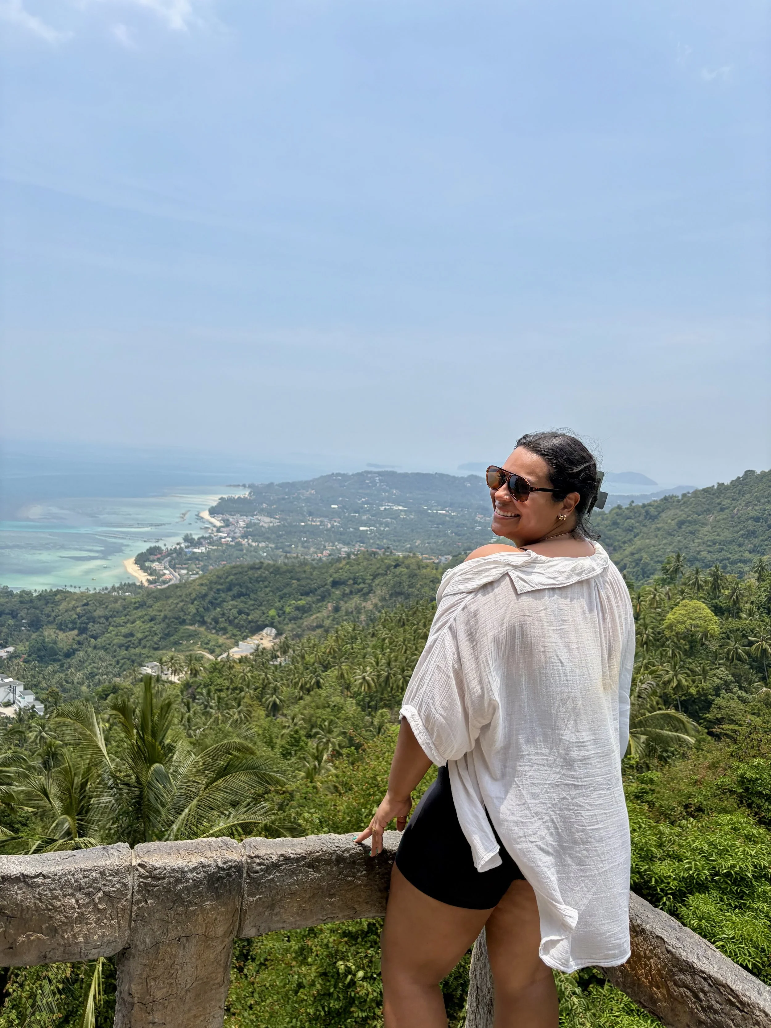 A woman wearing sunglasses and a white shirt standing on a stone railing overlooking a lush green landscape with trees, water, and distant islands under a blue sky with some clouds.