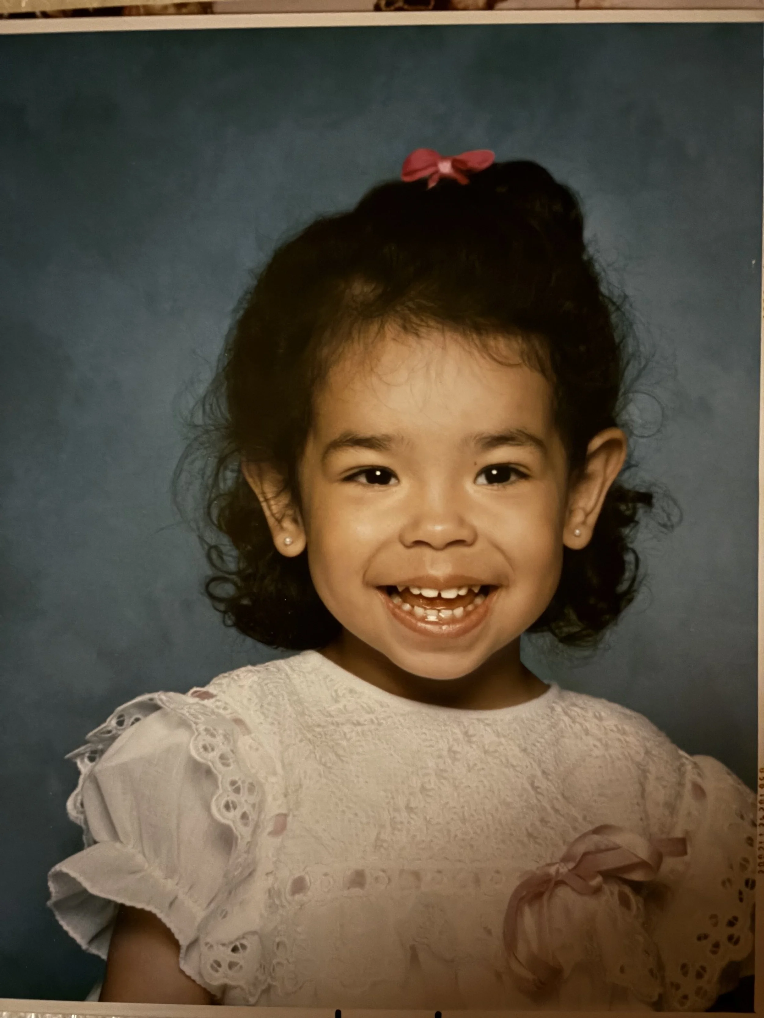 A young girl with dark curly hair, a pink bow clip, earrings, wearing a cream-colored lace dress, standing in front of a blue background, smiling and showing her teeth.
