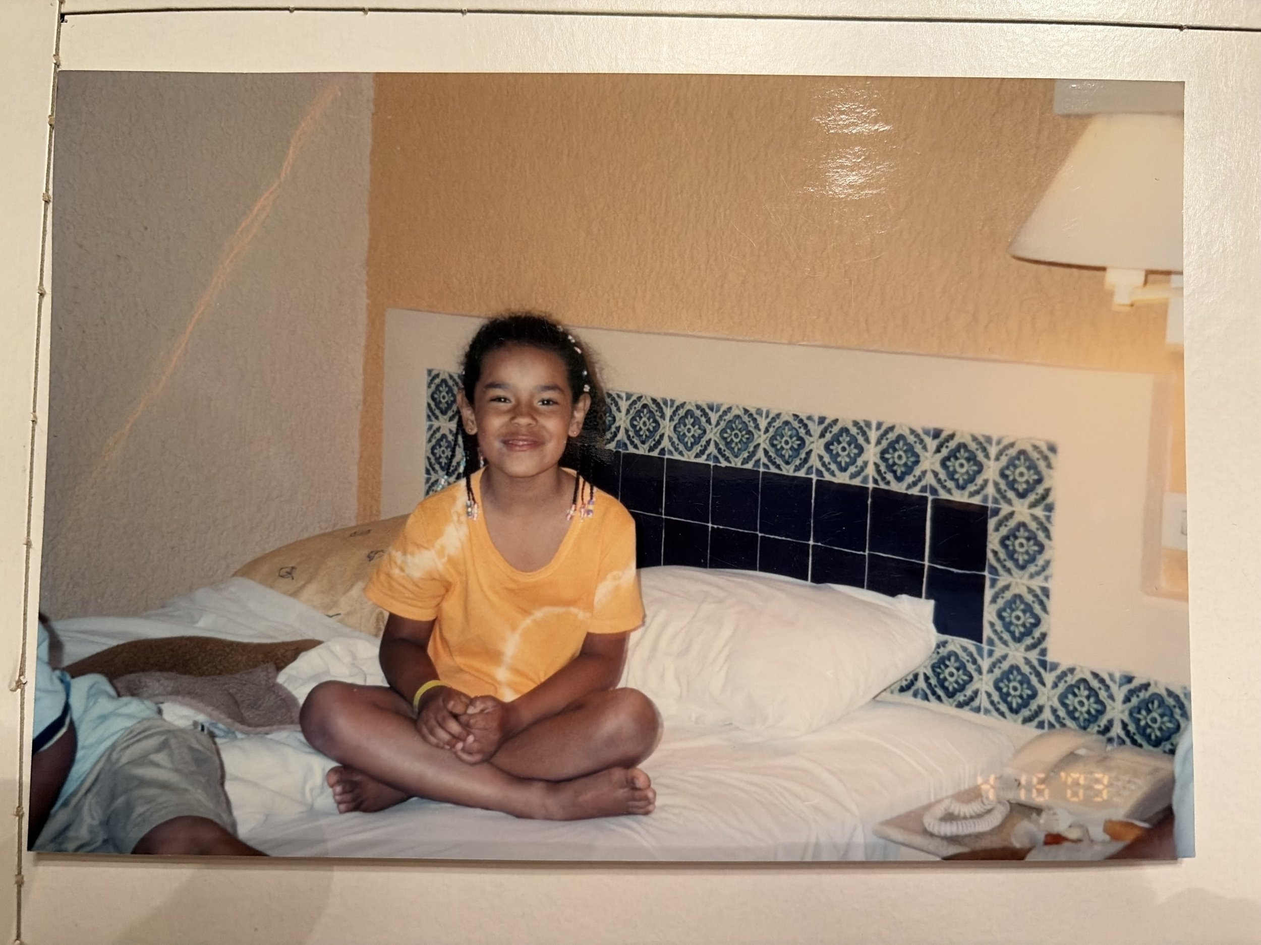 A young girl with a bright smile and dark hair tied back, sitting cross-legged on a bed with white sheets. She is wearing a yellow tie-dye shirt and jewelry with beads. The bedroom has warm-colored walls, a lampshade on the bedside table, and white and blue floral tiles on the headboard.