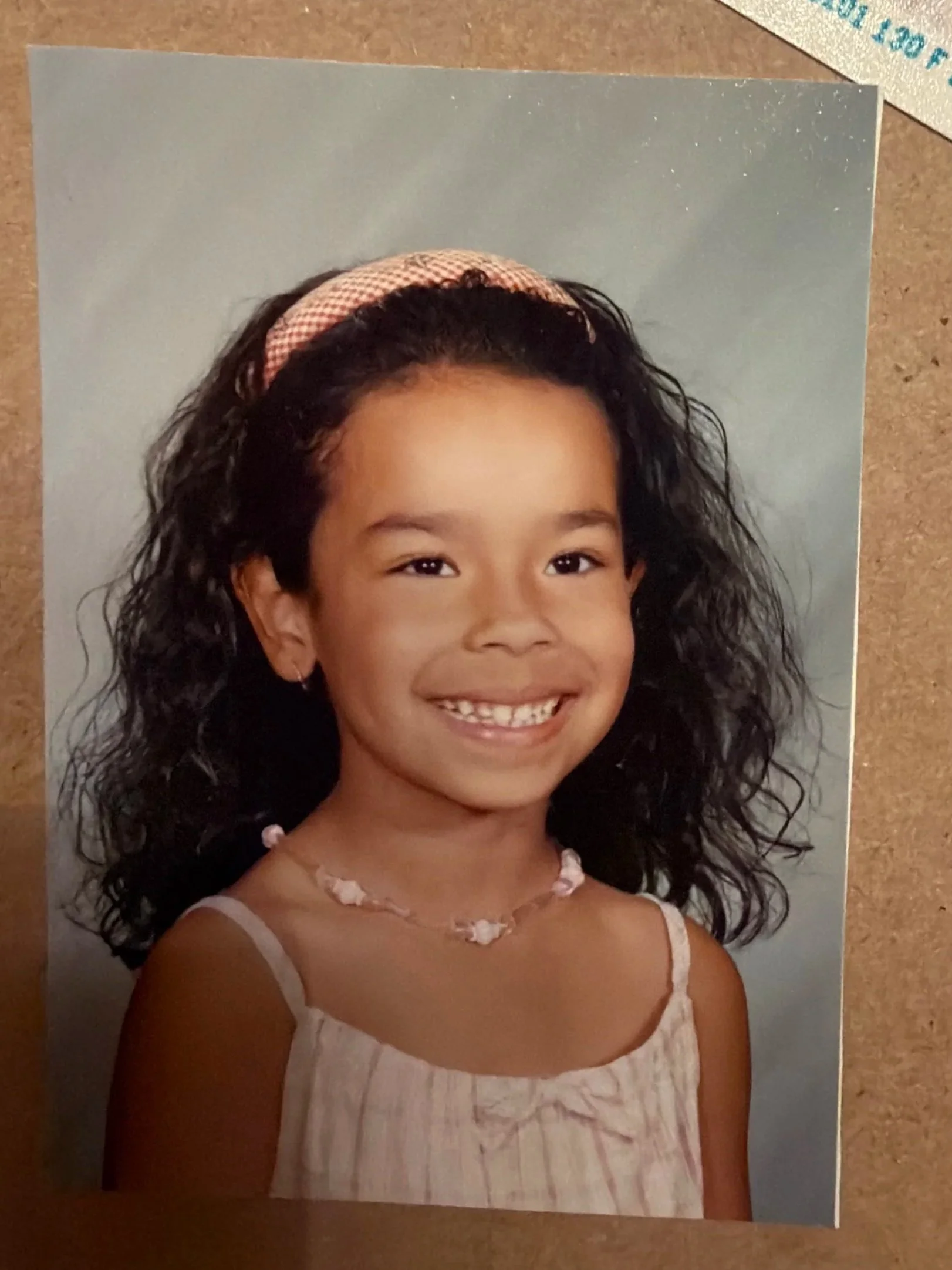 Photograph of a young girl with curly hair wearing a pink headband, smiling, in a white dress with pink accents and a beaded necklace, against a cloudy sky background.