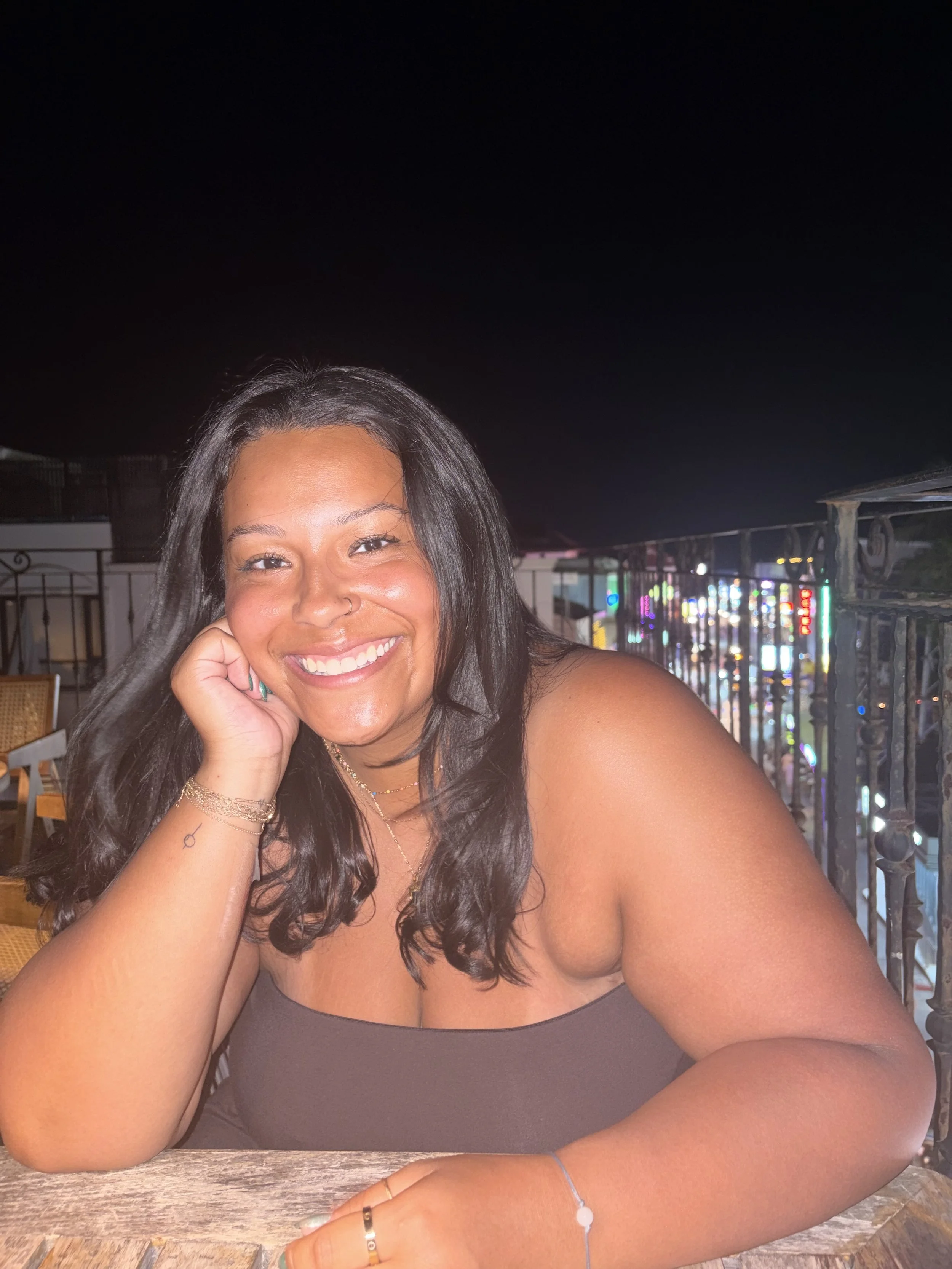 Close-up of a smiling woman with dark hair, resting her head on her hand, sitting at a wooden table at night, with city lights in the background.