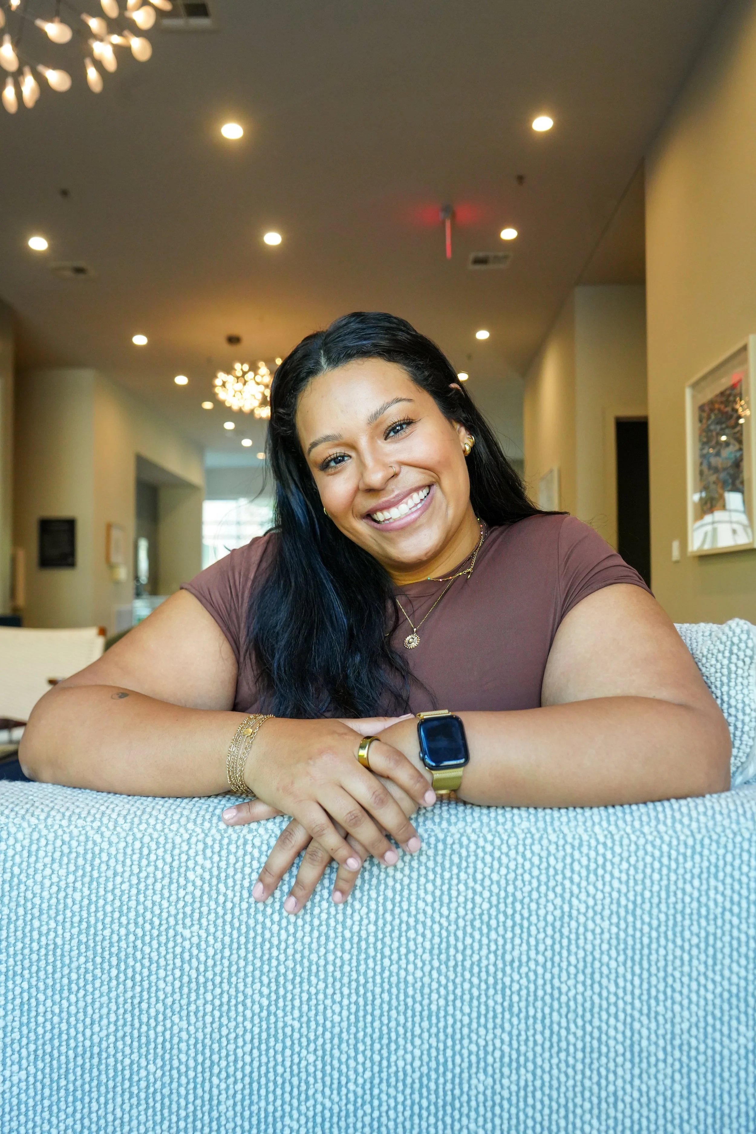 Smiling woman with long dark hair wearing a maroon top, gold jewelry, and an Apple Watch, sitting at a table in a brightly lit, modern interior.