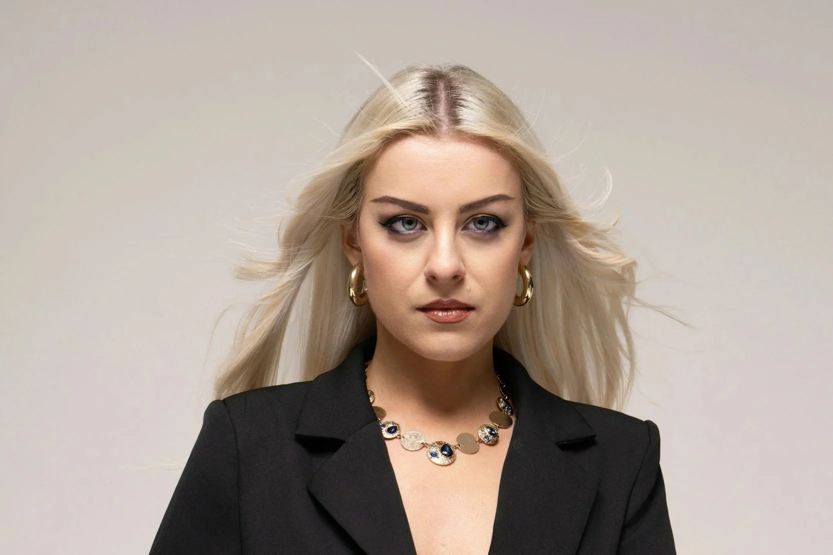 A smiling woman with natural curly hair, wearing glasses, earrings, and layered necklaces, in an indoor setting.
