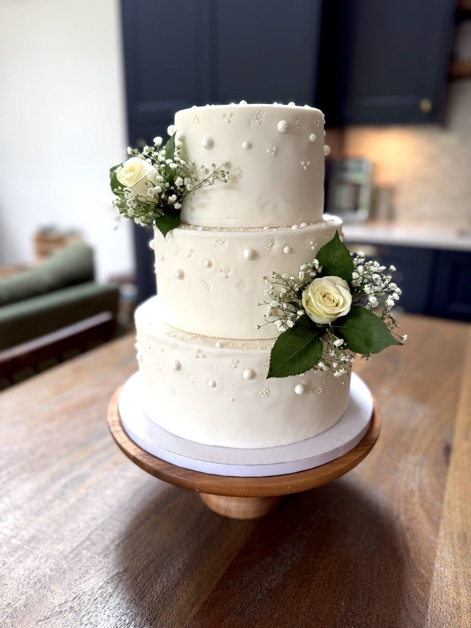 Three-tier white wedding cake decorated with small white pearls and fresh white roses with greenery, placed on a wooden cake stand.