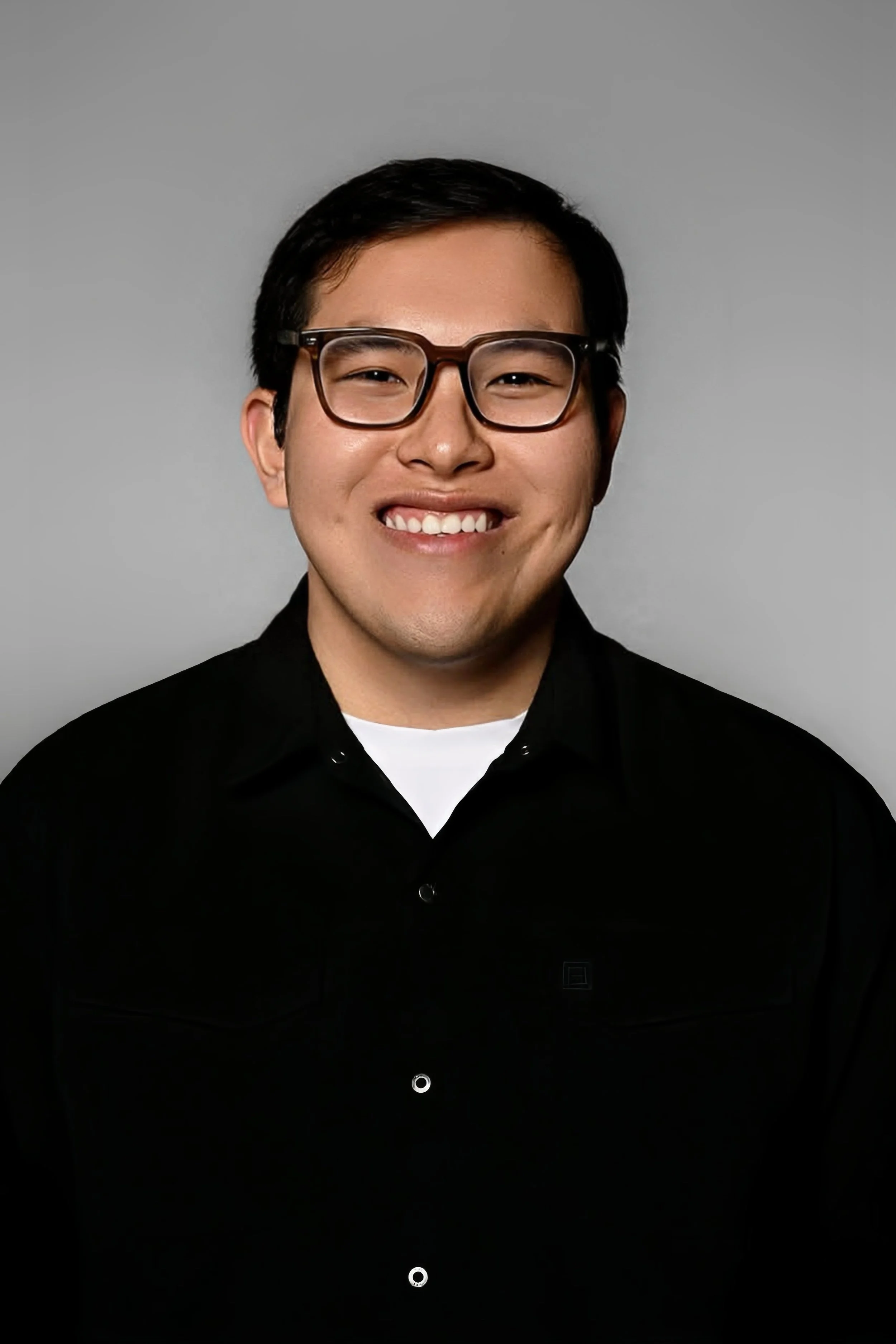 A smiling young man wearing glasses and a black button-up shirt against a plain grey background.