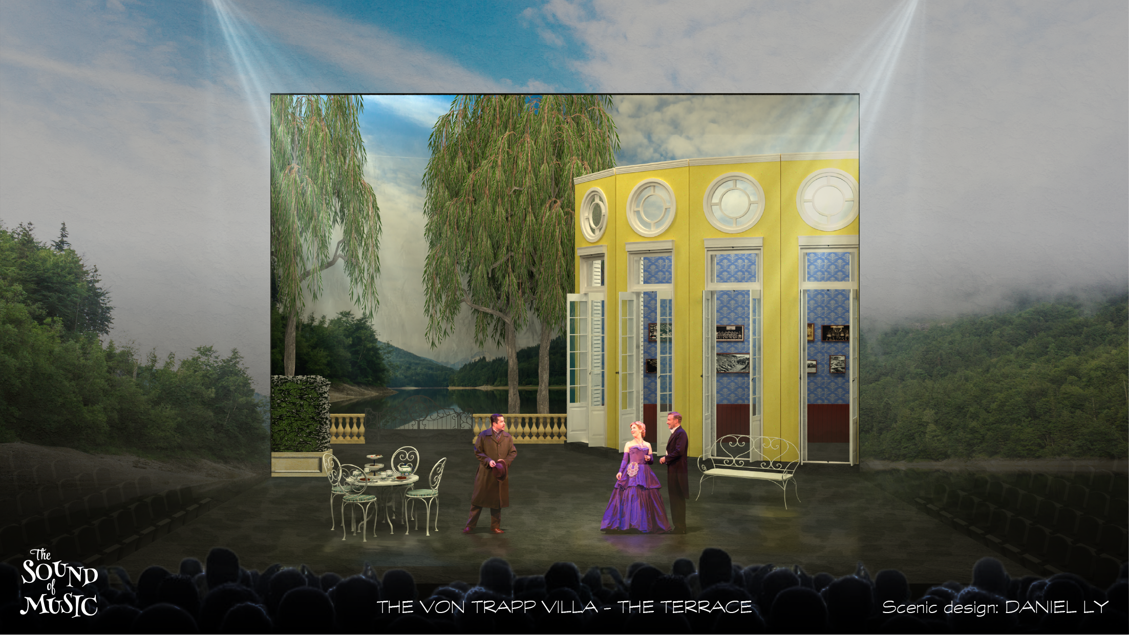 Scene from a theatrical performance set outdoors with trees and a cloudy sky backdrop. The stage features a large yellow mansion facade with multiple windows, a decorative railing, and outdoor furniture, including a round table with chairs. Four acto