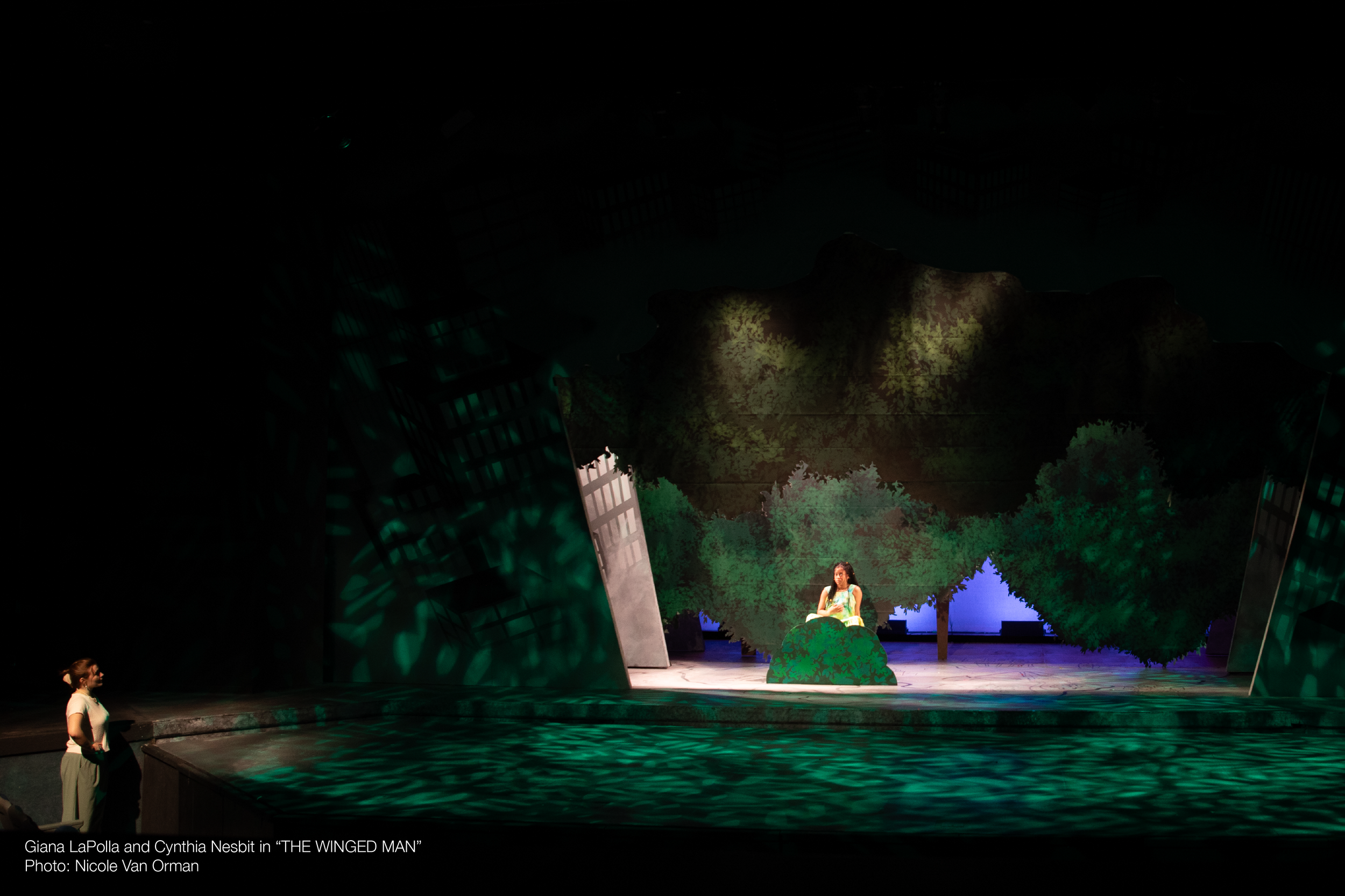 Stage scene from 'The Winged Man' featuring Giana LaPolla and Cynthia Nesbit, with stage scenery resembling trees and a cityscape background, and stage lighting creating a dark atmosphere.