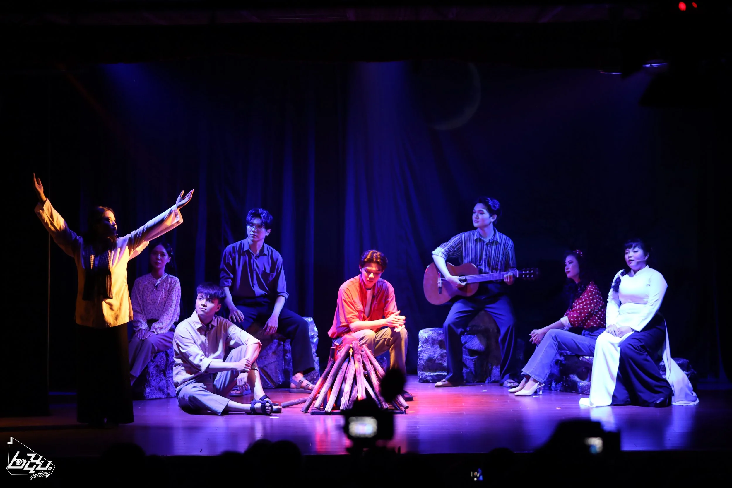 A group of young people performing on stage with a woman standing and gesturing with her arms. One person is playing a guitar, and there is a campfire prop in the center. The stage is lit with blue and purple lights, and dark curtains are in the back