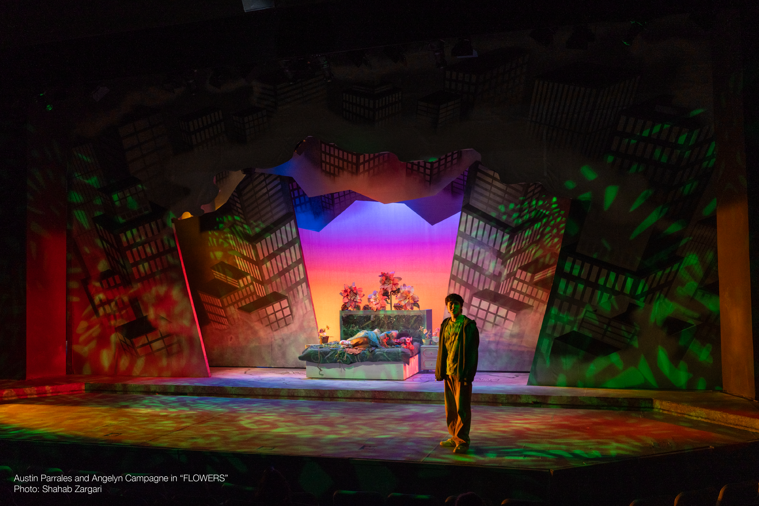 Stage scene from a theatrical production titled 'FLOWERS' featuring a set with geometric building-like structures, colorful lights, and a person standing near a bed with stuffed animals and a flower arrangement, with fog effects.