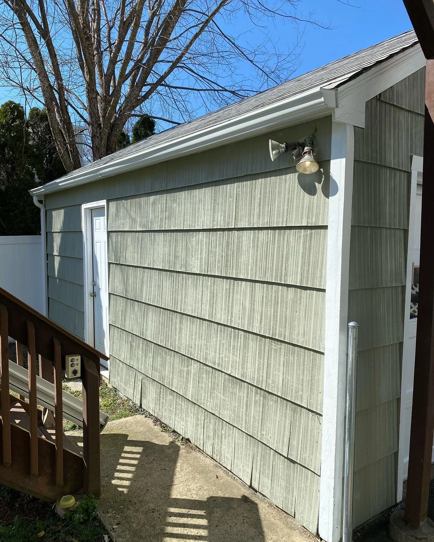A side view of a small, light green shed with horizontal siding, a white door, and a sloped roof. A twin outdoor security light is mounted on the corner of the shed. In the foreground, part of a wooden deck with railing and shadows is visible.