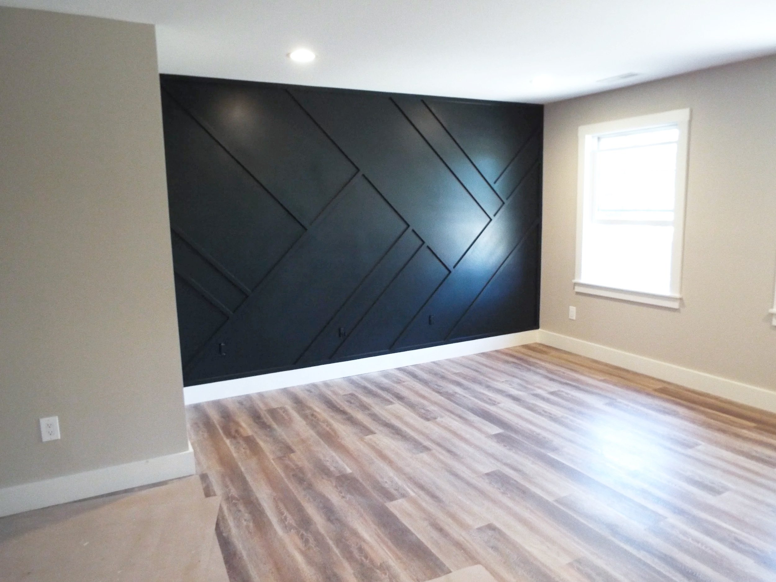 Empty room with a black accent wall featuring diagonal paneling, light wood flooring, and a window with white trim.