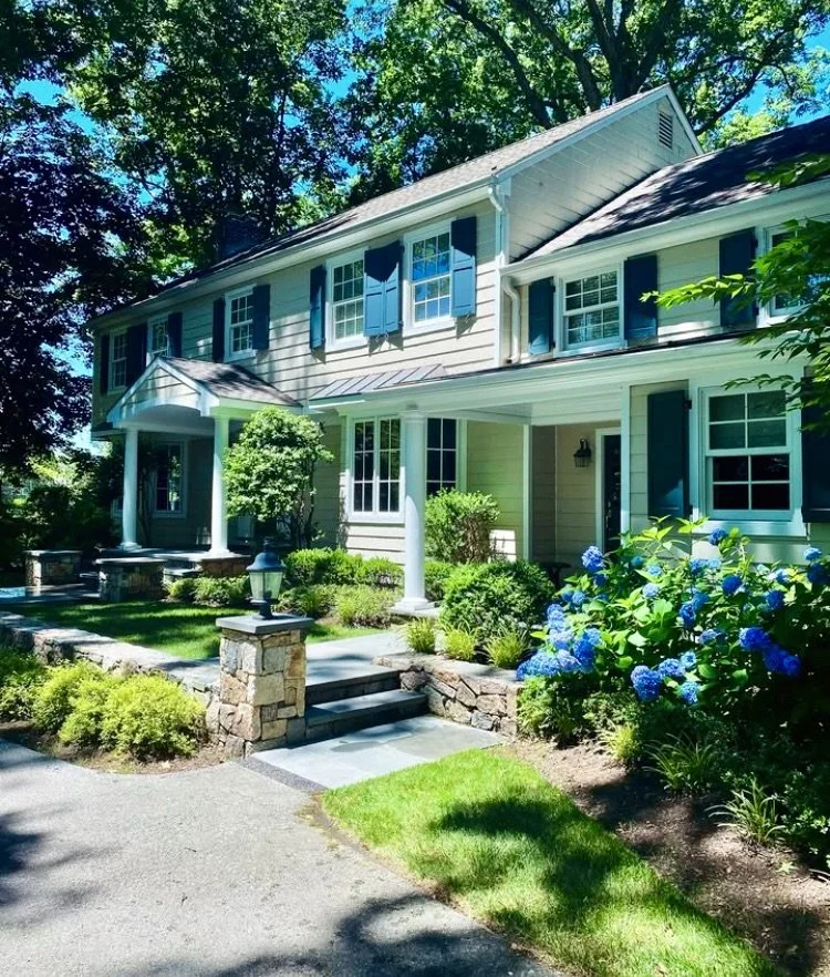 A large two-story house with beige siding, blue shutters, and a covered front porch surrounded by lush green landscaping and blooming blue hydrangeas.
