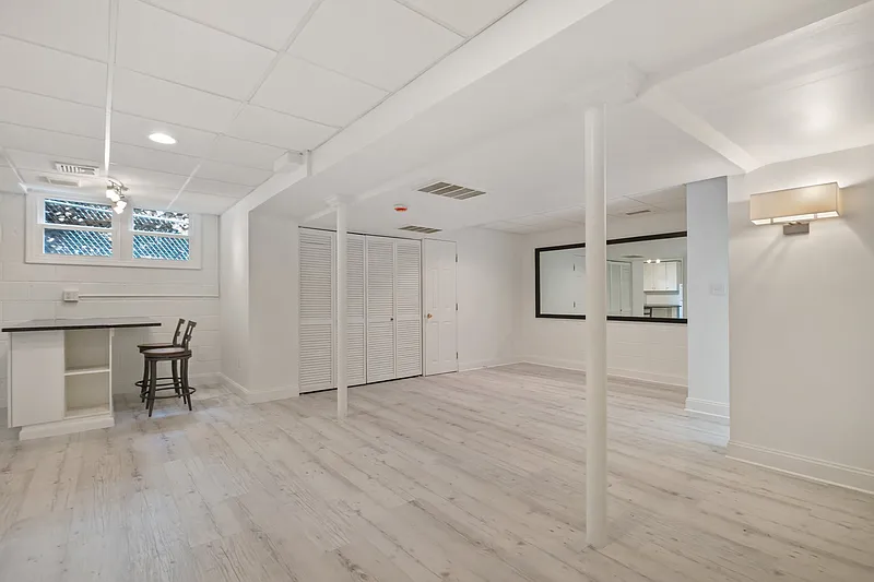 Empty room with white walls, light-colored wood flooring, a small breakfast bar area with two chairs, windows, a large mirror, and a ceiling light fixture.